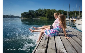 Zwei junge Mädchen sitzen auf einem Steg und planschen mit den Füßen im Wasser